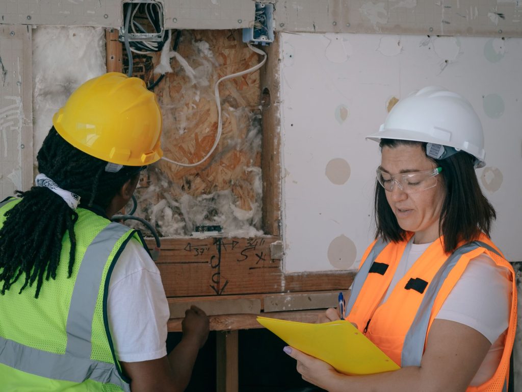 Two inspectors in safety gear are examining and documenting damage inside an exposed wall.