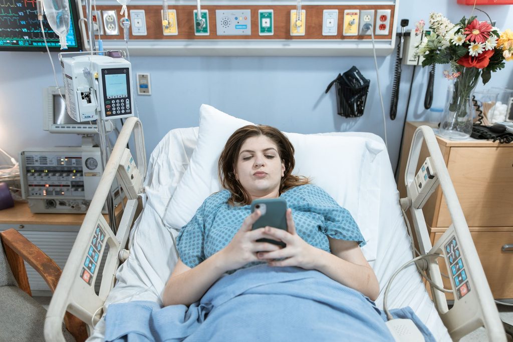 A female patient using her phone while lying on a hospital bed.